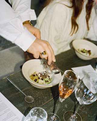 A person grates cheese over a bowl of pasta at a rustic table, whilst another person in a cream jumper sits nearby. There are glasses and a menu on the table.