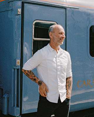 A man with tattoos wearing a white shirt stands and smiles in front of a blue train carriage on a sunny day.
