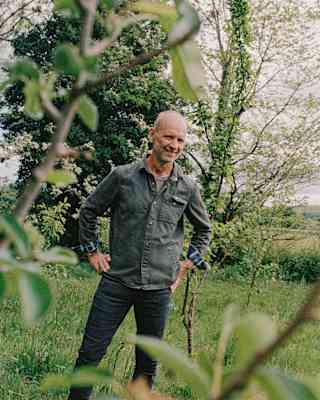 A smiling bald man in a denim jacket stands outdoors in a grassy area with trees and greenery in the background, partly framed by tree branches in the foreground.