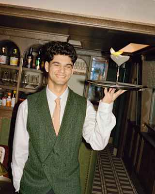 A smiling waiter in a green waistcoat, white shirt, and orange tie stands indoors holding a tray with a cocktail garnished with a lemon slice. Shelves with bottles are visible in the background.
