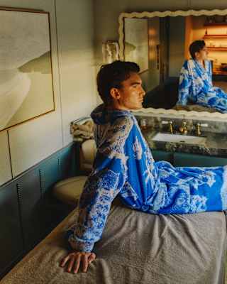 A man wearing a blue and white patterned dressing gown sits on a bed in a stylish room, looking to the side. His reflection is visible in a large mirror above a basin behind him.