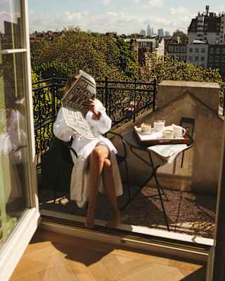 A female guest in a white bath robe reads a newspaper at a table set for morning coffee on a balcony with big views.