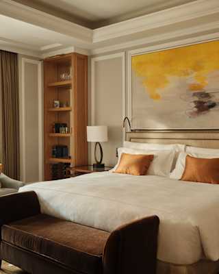 A yellow and grey abstract artwork hangs above a white Junior Suite bed with bronze cushions and a brown velvet bed bench.