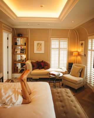 A guest's legs are seen on a bed in a view of mellow suite with backlit cornice, which glows above a chaise long and armchair.