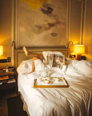 A tea-tray rests on a bed where a robed guest lies on top of the rumpled duvet, holding a newspaper in front of their face.
