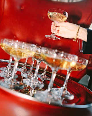 A hand lifts as glass of Champagne from a tray full of filled glasses, set on a red leather seat, seen in an angled close-up.
