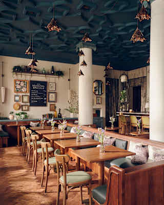 A dining room with natural tones, two white supporting pillars, teal ceiling and a long wooden banquette lined with tables.