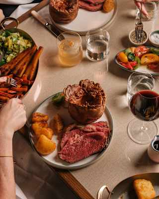 A guest reaches for carrots to add to a sumptuous plate of roast beef, potatoes and Yorkshire pudding, served with sides and wine.