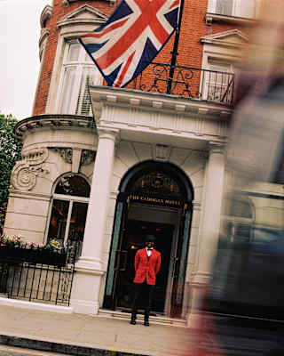 Tilted view across a busy road of a doorman in a red jacket with black trousers and bow tie outside the hotel entrance.