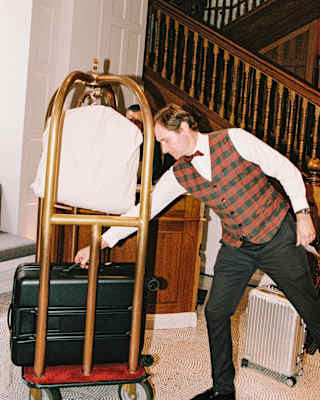A hotel porter in a tartan waistcoat, white shirt and black trousers reaches for a case on a luggage trolley in the lobby.