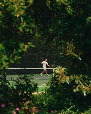 View through trees and shrubs of a tennis court, where a man in whites on the other side of the net reaches to hit a ball.