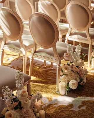 Two cream aisle bouquets and votive candles sit at the base of beige end-row chairs in a sunny ceremony room, seen close-up.