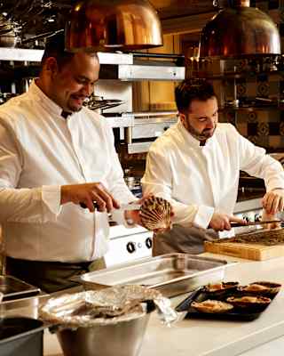 Wearing whites, Chef Michael Turner shucks a scallop over a tray as another chef prepares food next to him at the worktop.