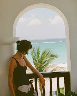 Through a balcony arch, a woman with dark hair in a black top gazes over a wind-blown palm at a white beach and sapphire sea.