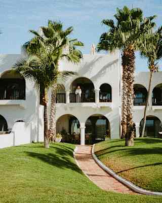 View from the garden to a white, Moroccan-style, two-level villa with arched colonnade balcony and terrace, flanked by palms.