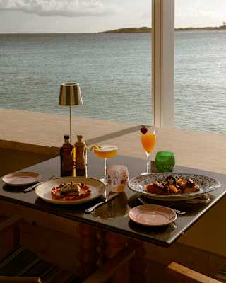 A table set for two with plates of food, drinks, and a lamp overlooks a calm sea through a large window, with sunlight reflecting on the water.