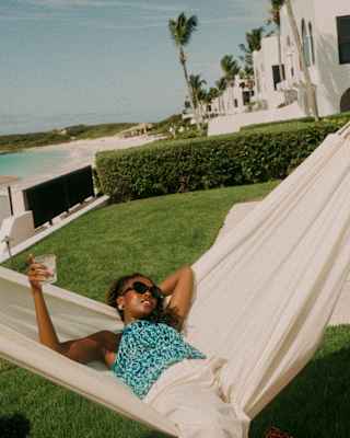A woman in shades and patterned blue top lies back in a white hammock with a drink, in a view over a private villa garden.