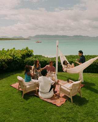 Guests chat in a lush green sea-view garden, with two on chairs, two on a blanket, one in a hammock and another standing.