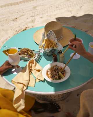 A couple enjoys light lunch with cocktails at a duck-egg blue table in the sand, with a sunhat on one side, seen from above.