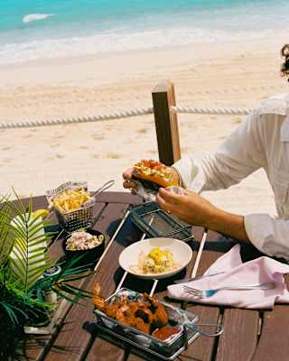 Looking across a vibrant Cap Shack lunch, enjoyed by a guest at a wooden table, to the dazzling white beach and sapphire sea.