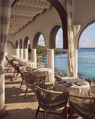 View through the seafront colonnade terrace at Pimms restaurant, where a row of six round tables for two are laid for lunch.