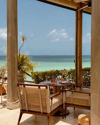 A table for two with rope-backed chairs claims a stunning tropical water view from the edge of a covered terrace lined with greenery.