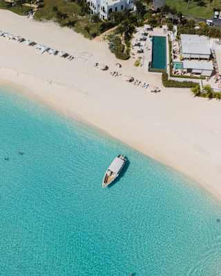 Bird's-eye view of the beach, where a boat floats in the shallows bordering white sands, with a glimpse of the resort behind.