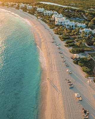Late sun sweeps over the soft sand beach, bordered by sparkling sea and white villas with lush gardens, seen from the air.