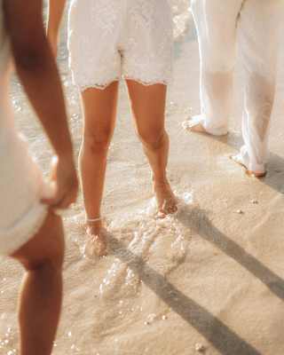 Four people, three in white skirts and one in trousers, play in the white water on the beach, seen close-up from waist down.