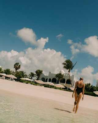 A person in a swimming costume walks on a sandy beach towards the camera, with clear shallow water in the foreground and a blue sky with scattered clouds above palm trees and a building in the background.