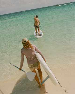 A woman carries a paddleboard into the clear sea, following a man who is already paddling ahead, seen from above and behind.