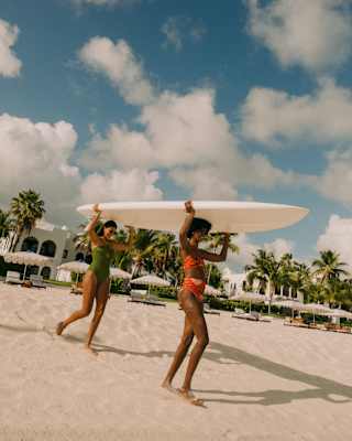 Two women in swimming costumes carry a surfboard on a sandy beach, with palm trees, blue sky, clouds, and white resort buildings in the background.