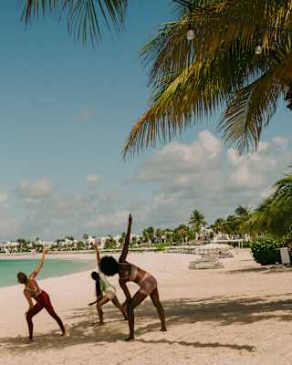 A group of five takes a yoga class on the beach, part-shaded by palm trees, with view across the waters of Maundays Bay.