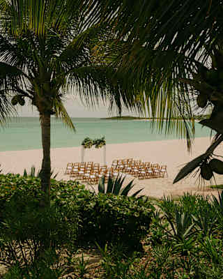 View from above through palm trees to a beach where two columns of chairs face a wedding arch with a sublime sea backdrop.