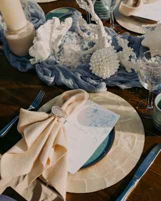 Close-up of a cream napkin on a capiz placemat at a wedding table with coral centrepieces arranged on wave-like blue cloth.