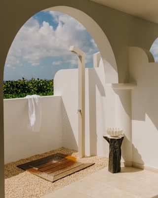 A white towel hangs over the wall of a sun-drenched outside shower with a wooden drainage mat, seen through an archway.