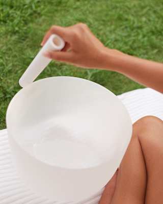 Close-up of a white singing bowl placed by the crossed legs of a woman who holds a sound mallet during a sound bath session.