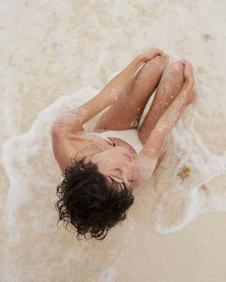 A woman with short dark hair sits on the beach, holding her knees, in the sea's white foam shallows, seen from above.