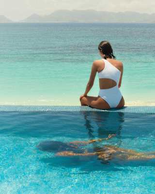 View over a pool where a guest swims underwater, and above her, a woman in white bathers sits on the edge facing the ocean.