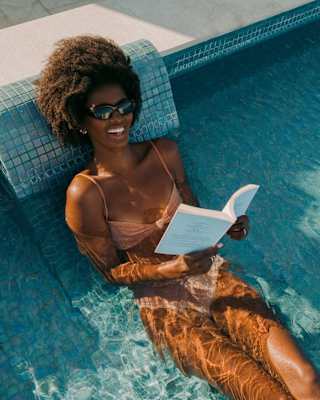 A woman in sunglasses relaxes on a poolside lounger in the water, wearing a swimming costume and reading a book under the sun.