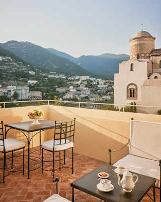A corner balcony table is set for coffee with views of Chiesa di San Giovanni del Toro and the higher slopes of Ravello