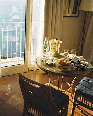 Sunlight pours through balcony doors with wide sea views, illuminating a round table, busy with glasses, wine and fruit.