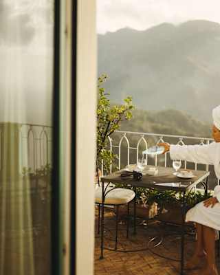 A woman in a robe and hair-towel pours a glass of water at a balcony table with misty hill views, glimpsed through a doorway.
