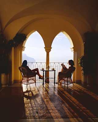 Sun through arches scatters shadows across Bar Caruso's terrace where two guests sit with drinks and sublime Amalfi views.