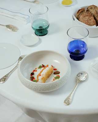 A white bowl with a gourmet dish, garnished with herbs and colourful toppings, sits on a white tablecloth. Nearby are silver spoons, blue and green glasses, a plate of bread, and a small dish with olive oil.
