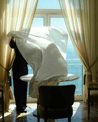 A white table-cloth obscures the face of the staff member who shakes it out as she lays a table set by a sea-view window.