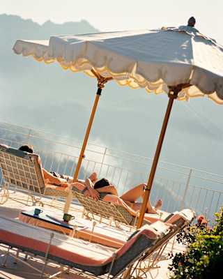 A man and woman recline on sun-loungers beneath two parasols, facing breathtaking views, seen from behind at an angle.