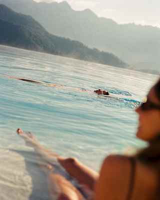 A man swims on his back in the pool, seen over the shoulder of a woman in soft-focus in an image that captures the hill views.