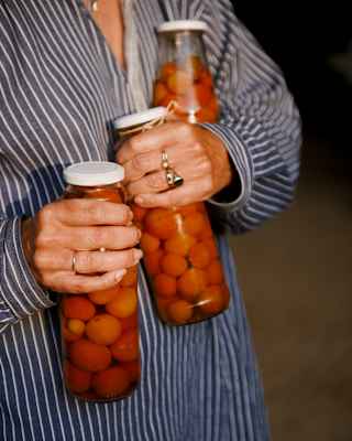 A woman in a striped shirt carries three tall jars of pickled cherry tomatoes, one in each hand and one under her left arm