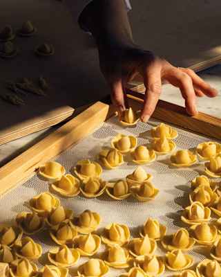 A hand delicately places a freshly-made tortellini pasta shape on a mesh tray with others, in a close-up of a pasta making class.